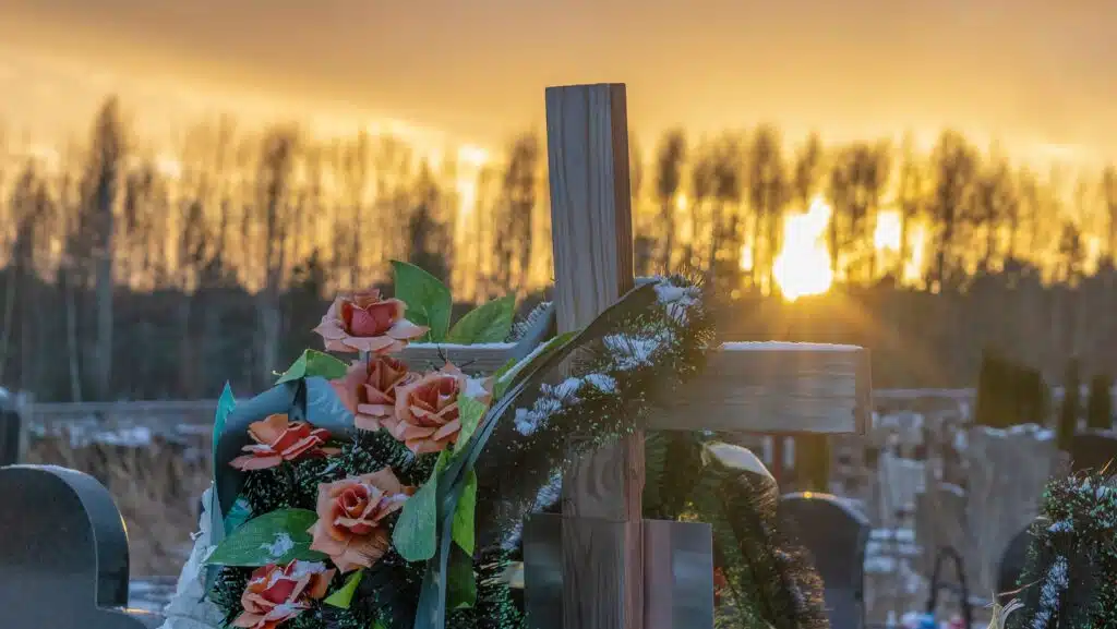 A wooden cross with a wreath over it in a cemetery at dawn.
