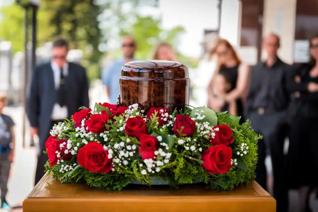 Funerary urn with ashes of dead and flowers at funeral. Burial urn decorated with flowers and people mourning in background at memorial service, sad and grieving last farewell to deceased person.