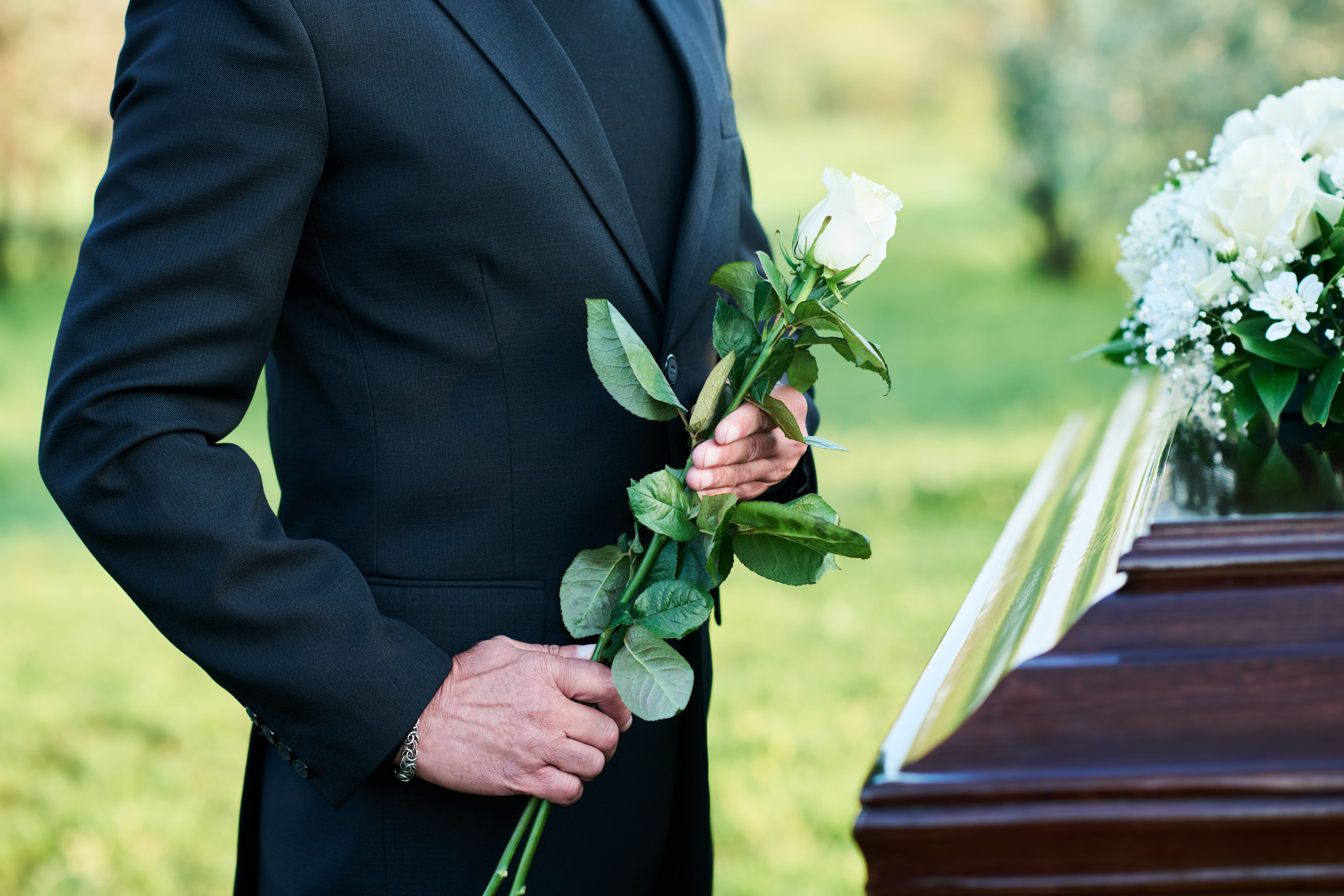 Man wearing traditional black suit at a funeral near a casket holding flowers